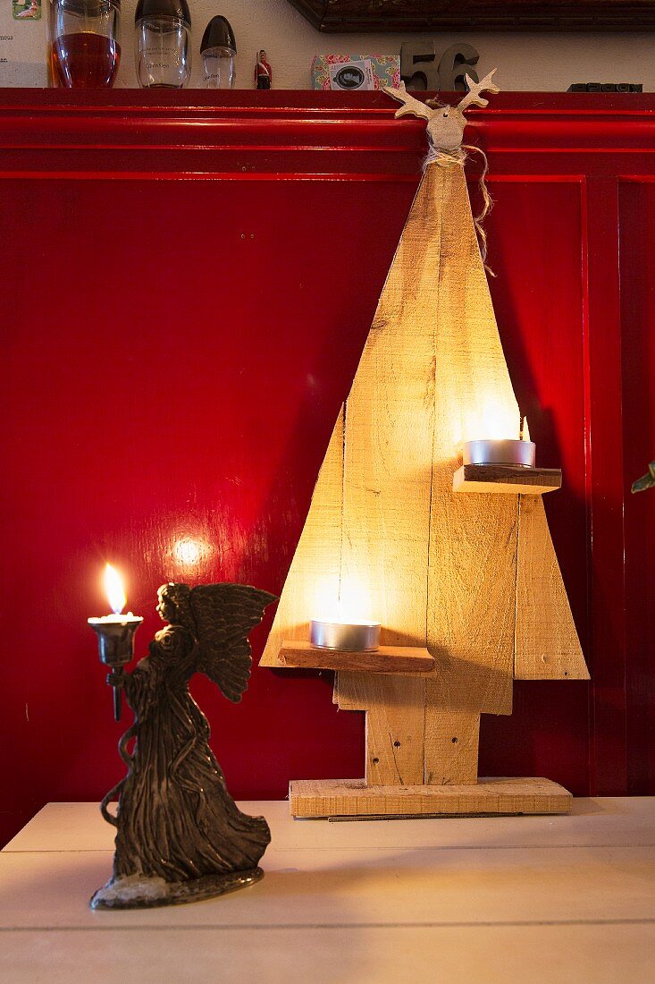 Ausgesägte Weihnachtsbäume mit Teelichtern und Engelsfigur mit Kerze vor roter Wandverkleidung