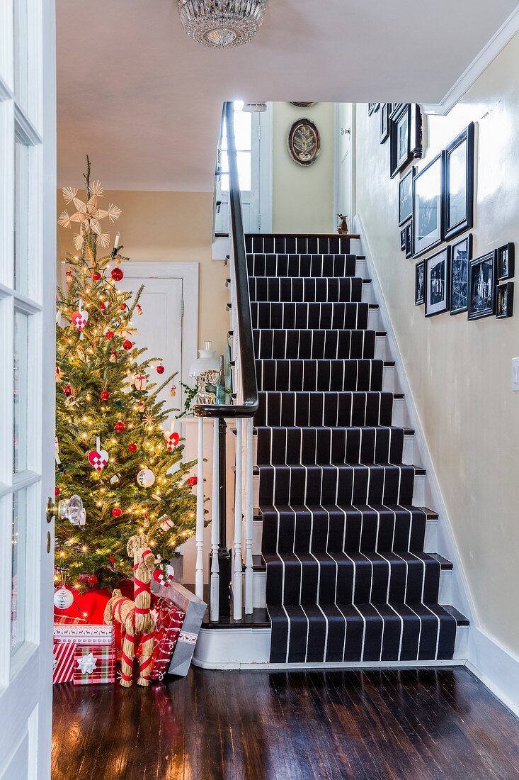 Geschenkpäckchen und Strohpferd unter rot-weiss geschmücktem Weihnachtsbaum neben Treppenaufgang in Landhausdiele