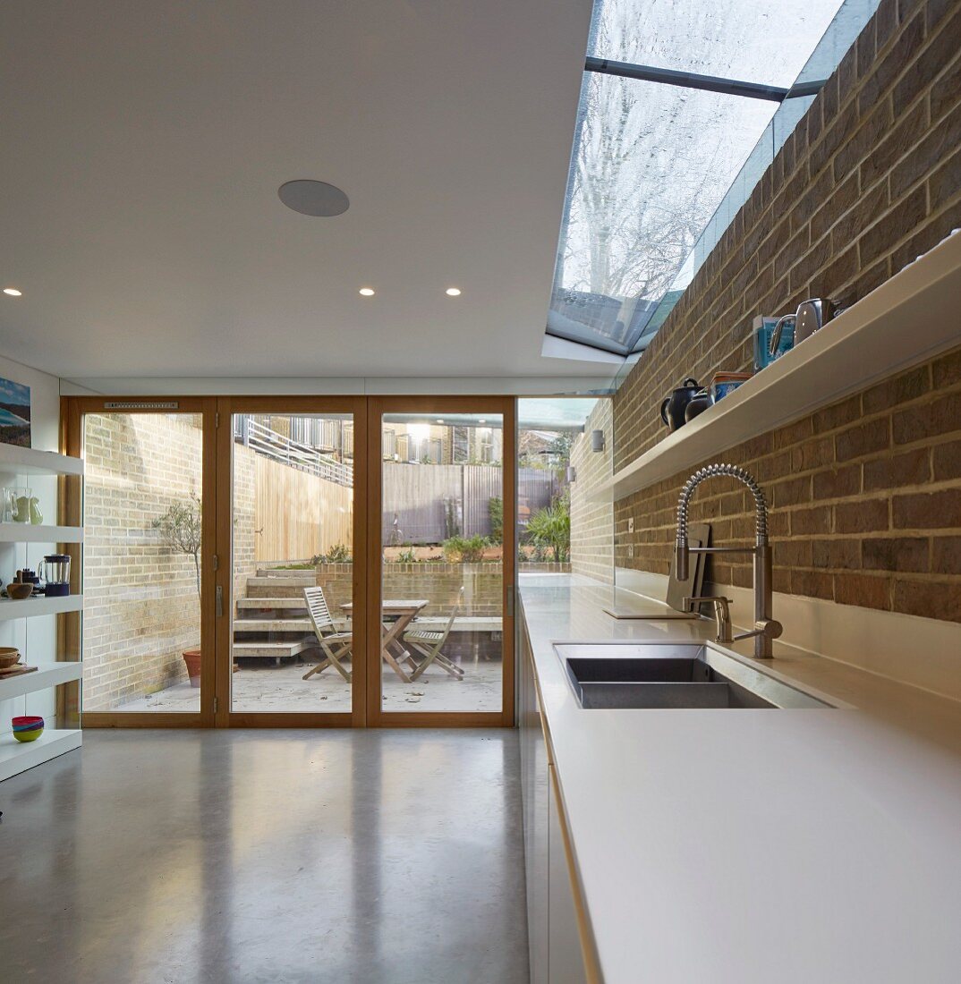 weiße Küchenarbeitsplatte in Designerküche vor Sichtmauerwerk, freier Terrassenblick mit Treppe und Gartenmauer