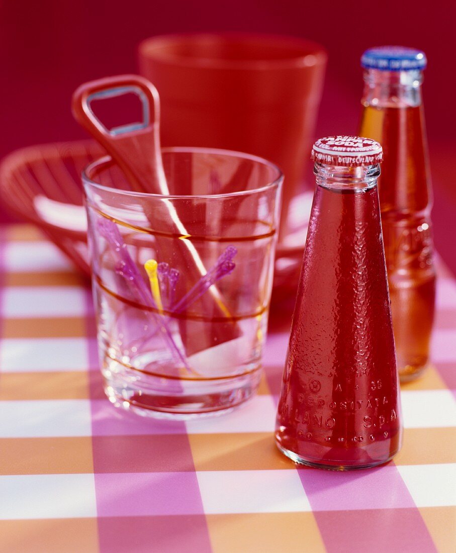 Small bottle of Campari and glass holding cocktail sticks and red bottle opener on pink, checked surface