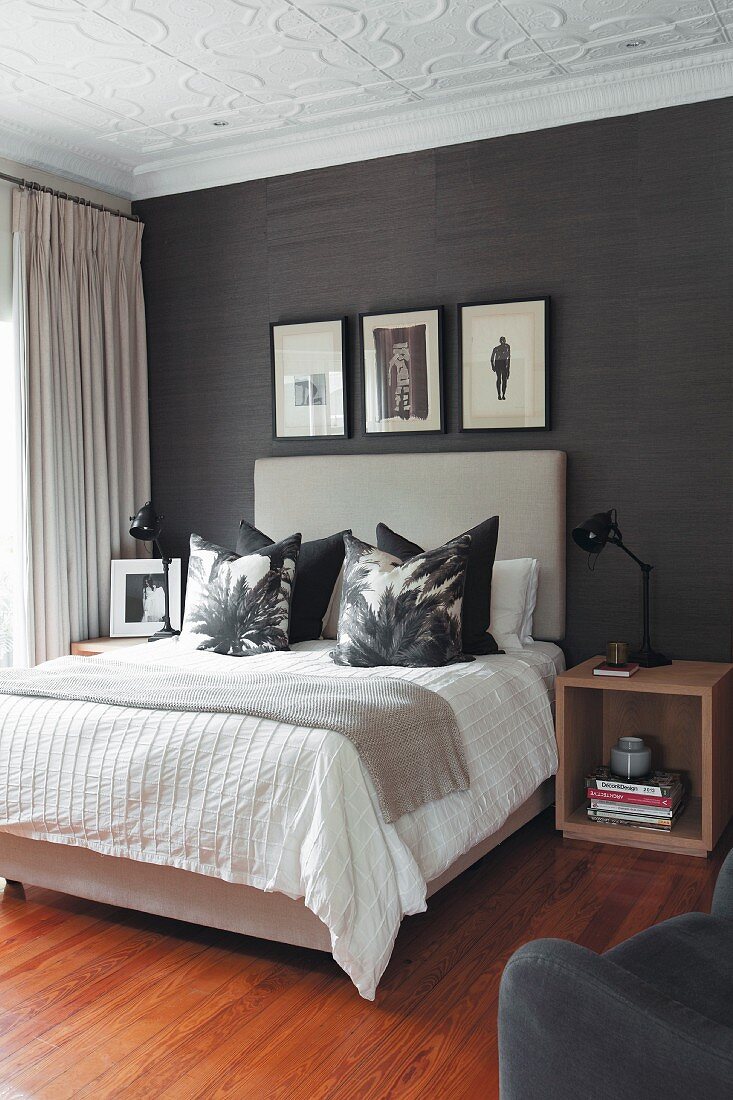 Scatter cushions arranged on double bed with tall, upholstered headboard against black wallpaper in elegant bedroom