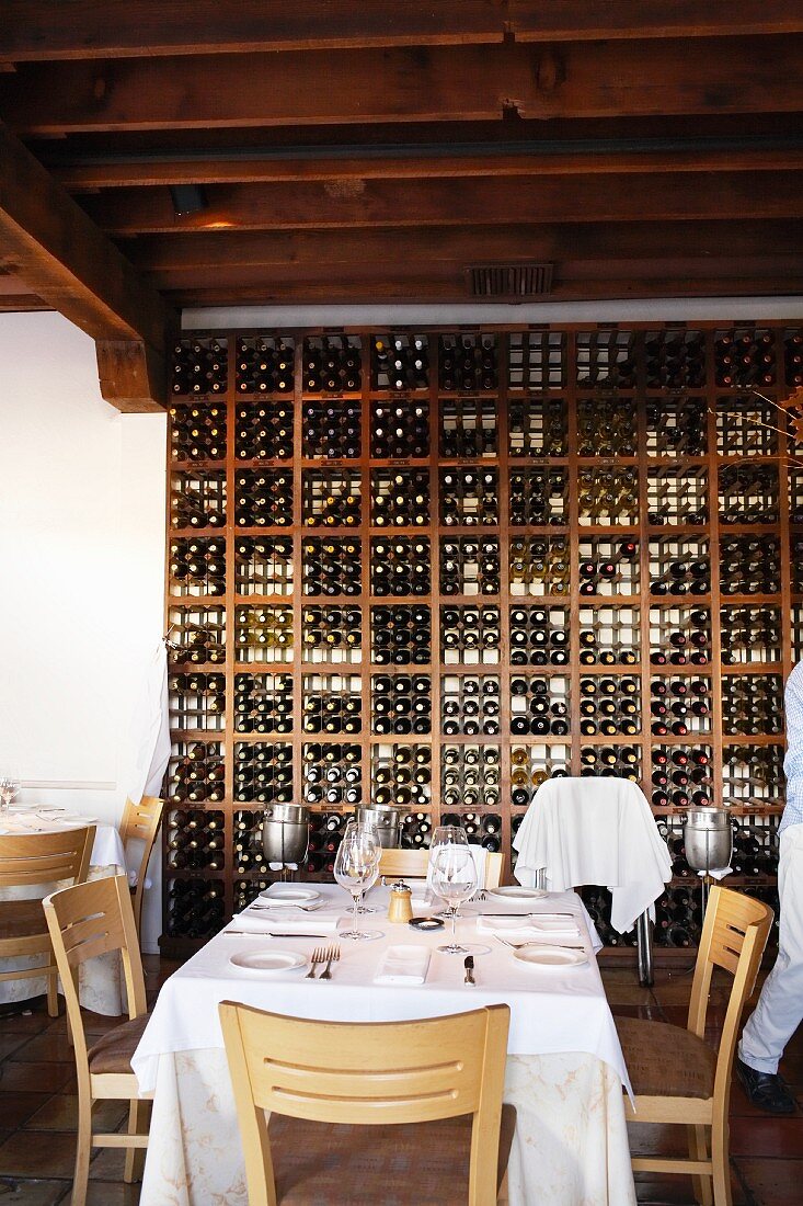 Weiß gedeckter Restauranttisch vor hohem Weinregal unter dunkler Balkendecke