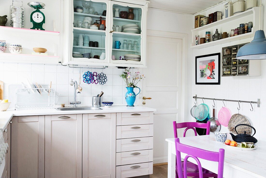 White-painted, L-shaped, country-house-style kitchen counter and wooden chairs painted purple in dining area in foreground