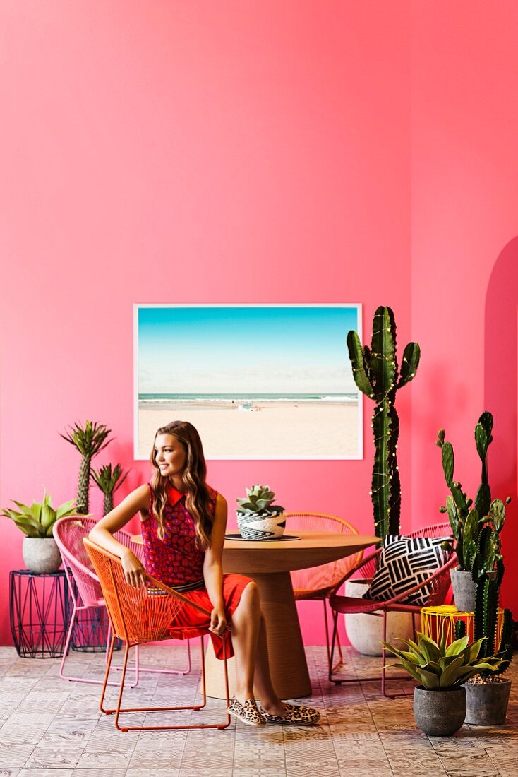 Retro-Urlaubsflair mit vielen Kakteen: Junge Frau sitzt auf Acapulco Chair am runden Tisch vor pinker Wand