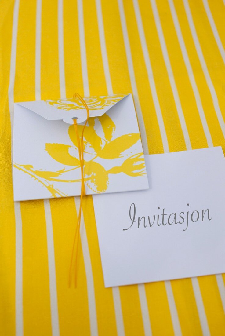 Einladungskarte mit gelbem Blütenmotiv auf weißem Kuvert auf gelb weiss gestreifter Unterlage