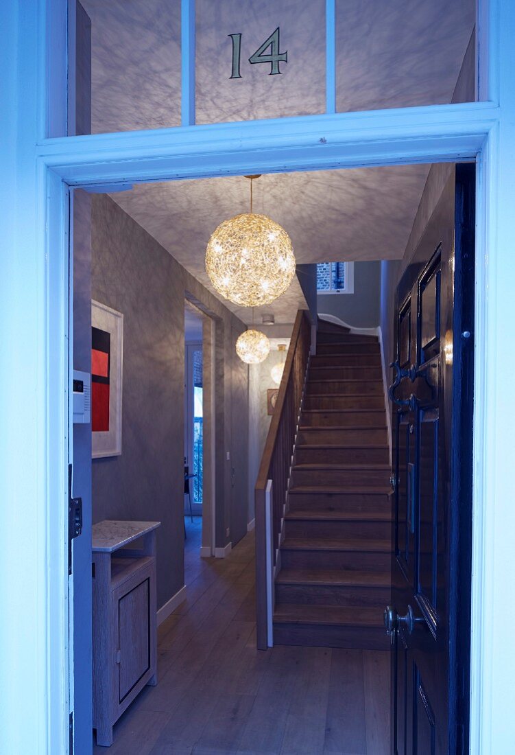 View through open front door into hallway with staircase and pattern of light and shade thrown by wicker lampshades