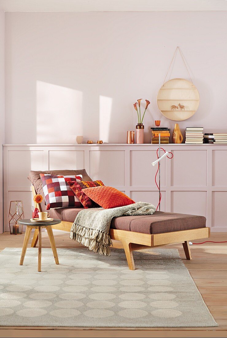 Gemütliche Tagesliege im Fiftiesstil mit Kissen und Tagesdecke, Beistelltisch auf gemustertem Teppich, halbhohe Holzwand und Wand in Pastellviolett