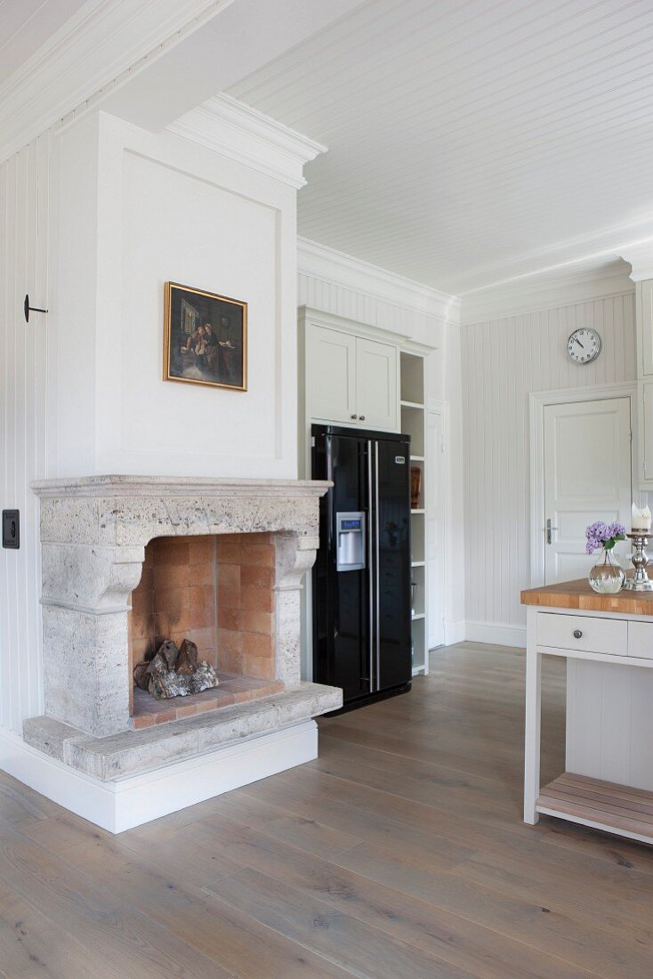 Open fireplace with stone surround next to modern, black fridge-freezer in white, wood-clad, country-house kitchen