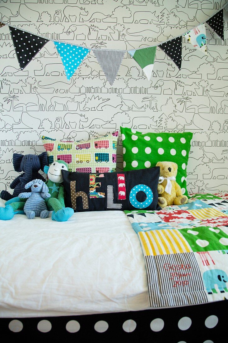 Wimpelgirlande über Kinderbett mit bunten Auflagen und Kuscheltieren, dahinter Tapete mit grafischem Tiermuster