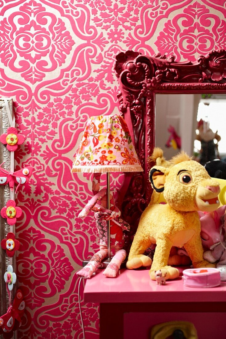 Soft toy, table lamp and framed mirror on pink table against wall with patterned wallpaper