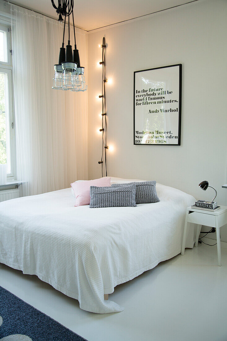 Scatter cushions and white bedspread on double bed and vertical string of fairy lights next to framed text artwork in bedroom