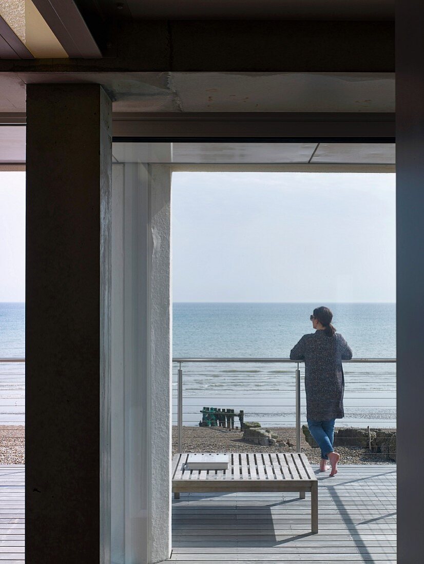 Blick vom Haus durch offene Terrassentür auf Frau an Brüstungsgeländer am Meeresstrand