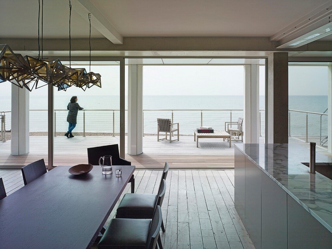View from dining area with dark table and upholstered chairs through open sliding glass wall to wooden terrace and sea