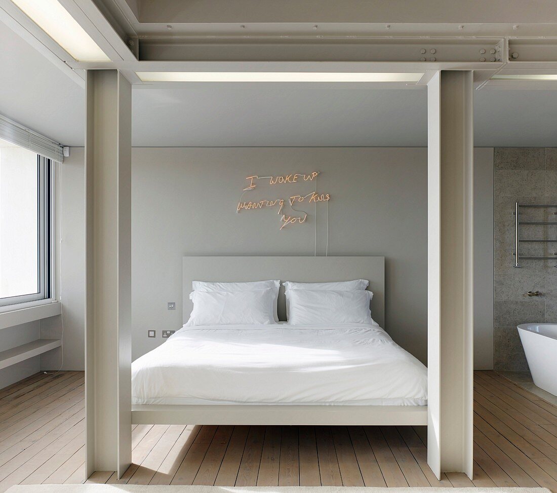 View between pale, steel-beam structure to double bed with white bed linen and illuminated lettering on wall