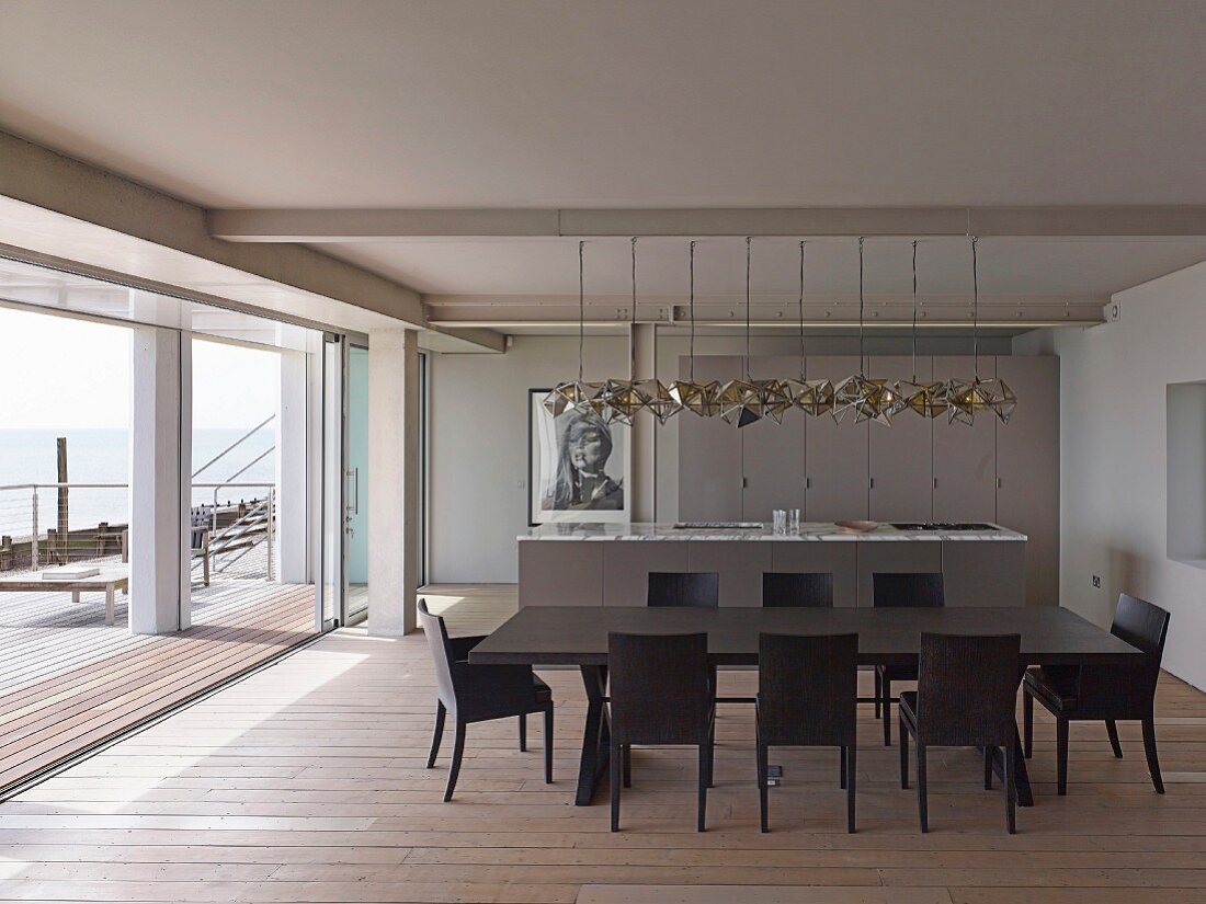 Dunkler Esstisch mit passenden Stühlen auf Dielenboden, Küchentheke und Designer Pendelleuchte in offenem Wohnraum mit Blick über Terrasse auf Meer