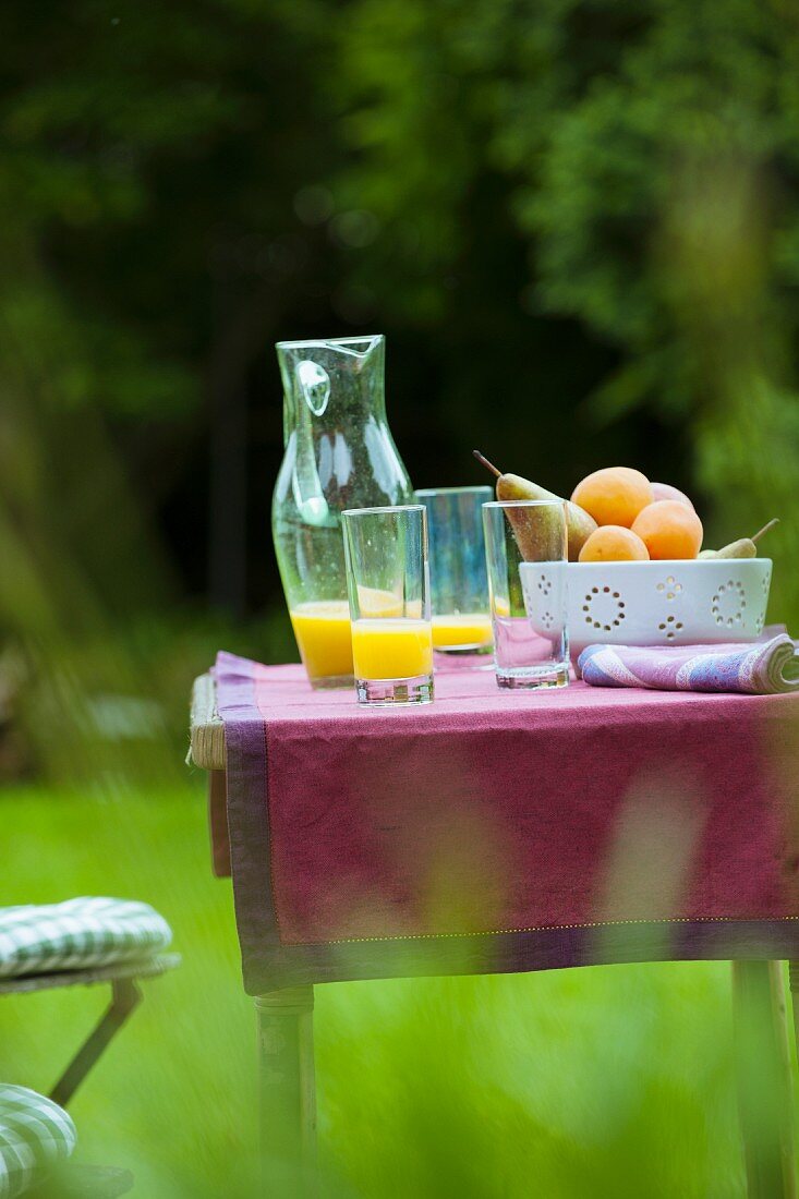 Erfrischungsgetränke und Obstschale auf Gartentisch mit violettrotem Tischläufer
