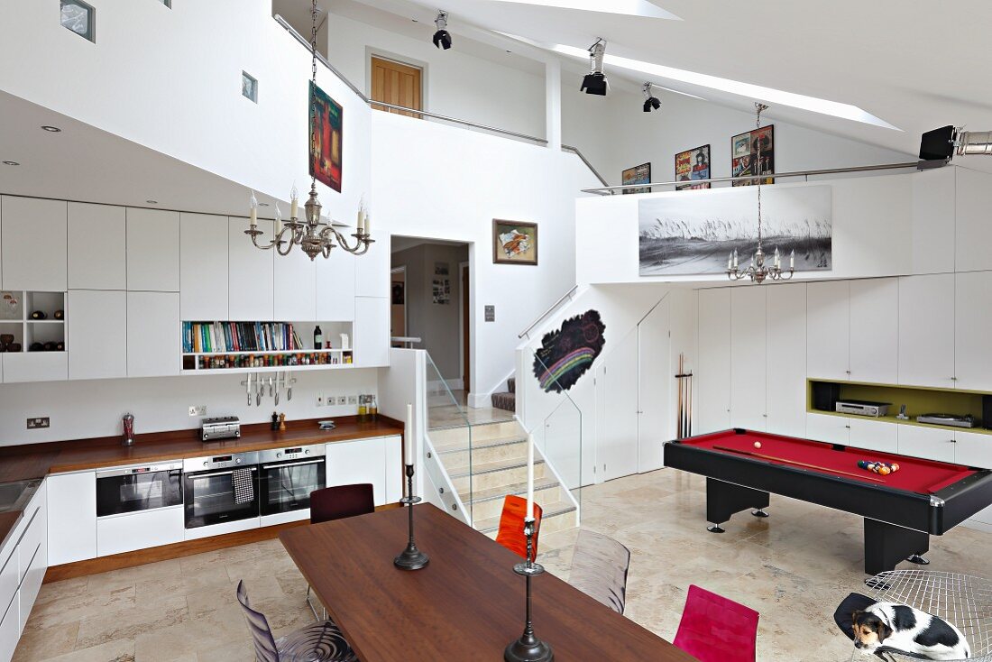View across dining table and through fitted kitchen to pool table in open-plan interior with encircling gallery