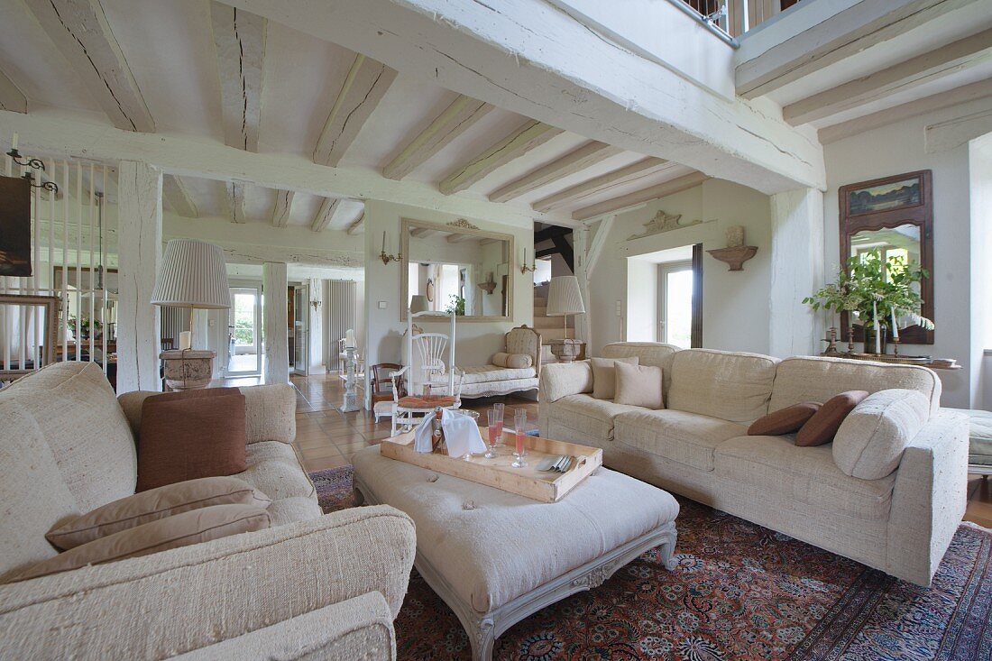 Pale sofa set and ottoman in open-plan living area of renovated country house with white-painted wood-beamed ceiling