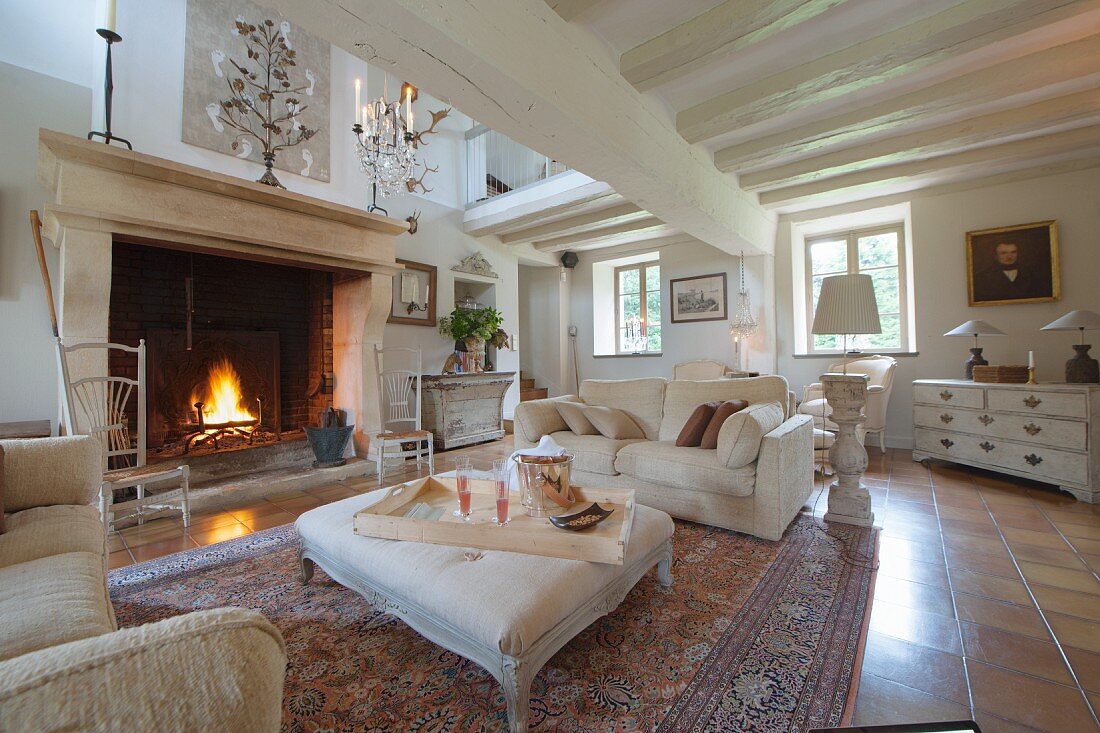 Pale sofa set and ottoman in front of fire in large fireplace in open-plan country-house interior