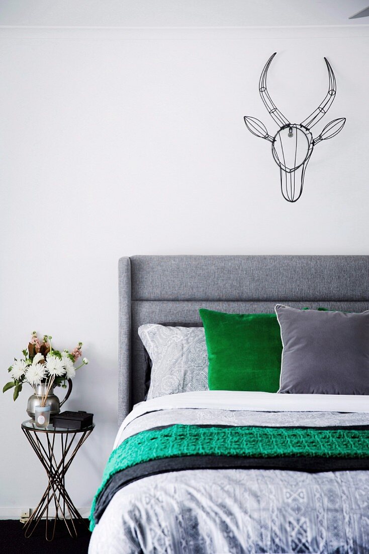 Bed with gray headboard, pillows and bed linen in gray and green, above wall decorative animal trophy made of black wire