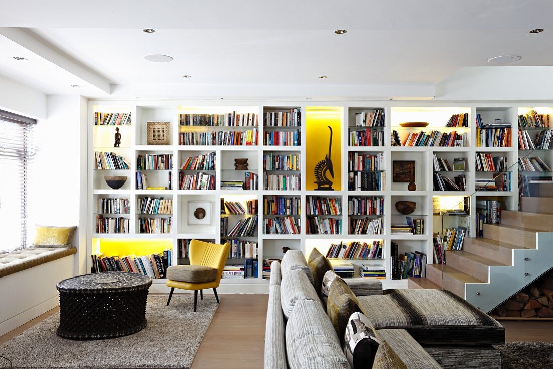 Offener Wohnraum mit Bücherwand teilweise beleuchtet, Sofa und Sessel mit Ethno-Tisch vor Fenster
