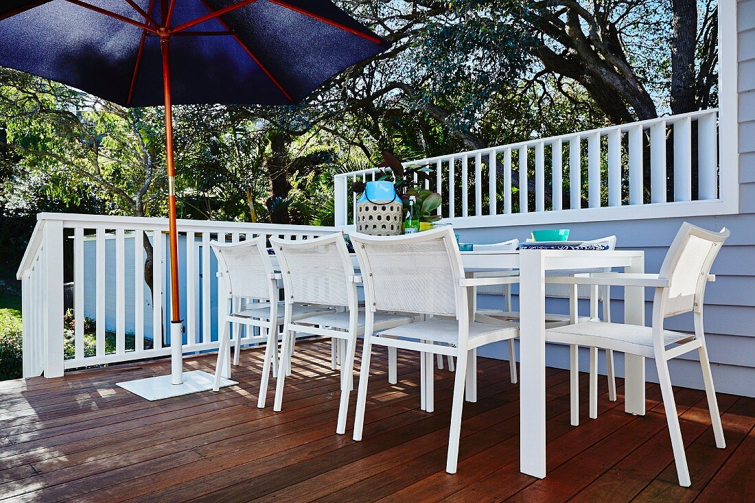 weiße Outdoor-Stühle und Tisch auf Terrasse, seitlich blauer Sonnenschirm vor weisser Holzbalustrade