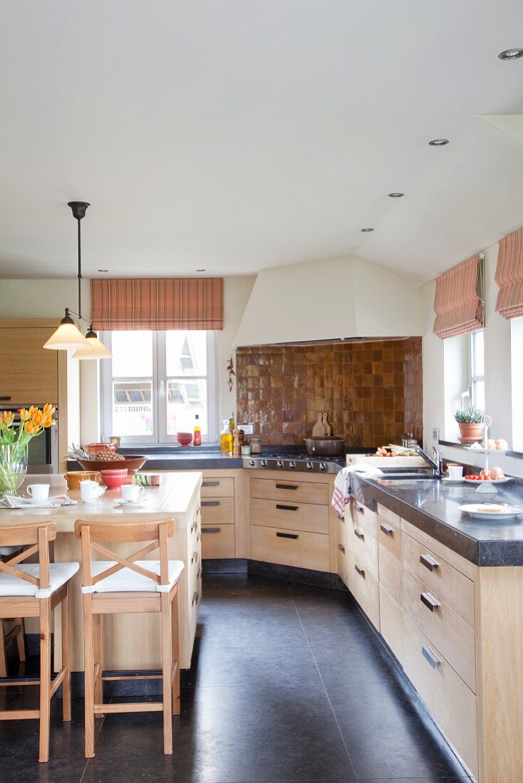 Elegante Landhausküche mit langer Küchenzeile, dunkle Stein Arbeitsplatte auf Unterschränken aus hellem Massivholz, seitlich Theke mit Barhockern
