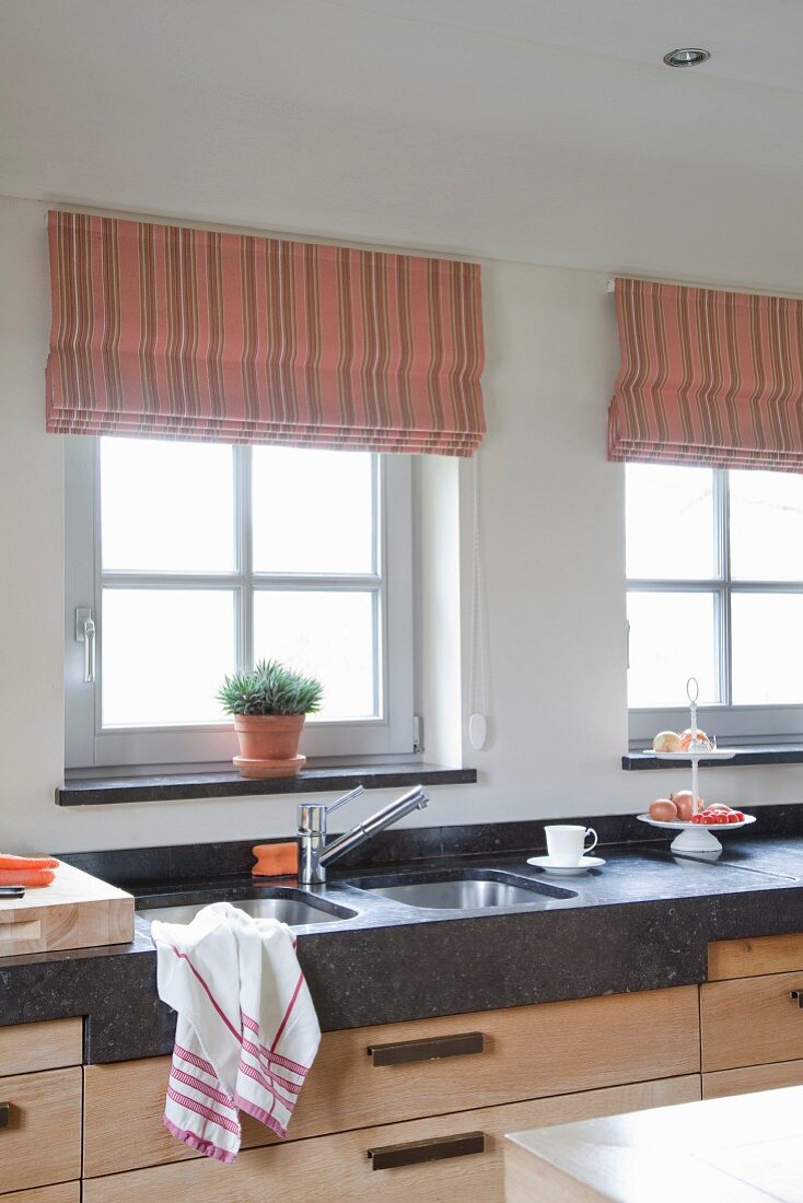 Elegante Küchenzeile mit dunkler Stein Arbeitsplatte, eingebaute Doppelspüle vor Fenster mit Raffrollo in pastellfarbenem Streifenmuster