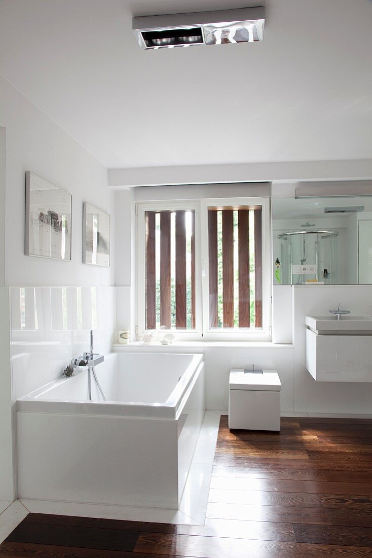 Badezimmerecke mit Fenster, weißer Wanne, WC und Waschbecken in kantigem, modernem Design dazu elegantes Edelholzparkett
