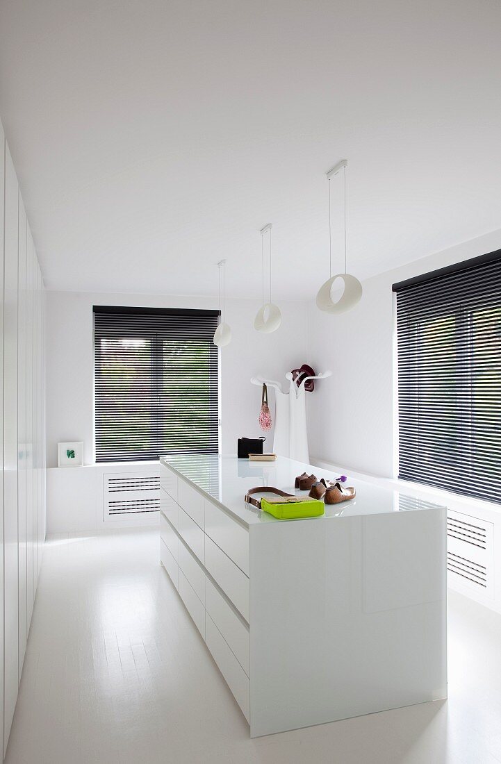 Central white chest of drawers, white designer lamps and black louvre blinds on window in minimalist dressing room