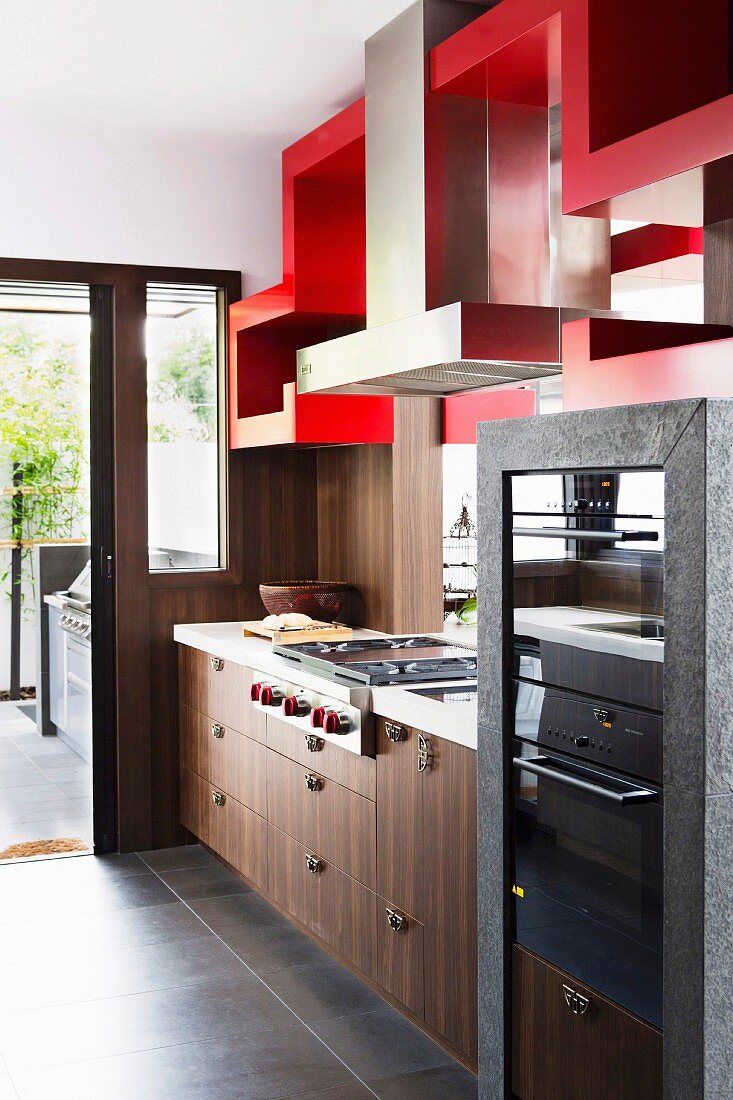 Küchenzeile mit edler Holzfront, oberhalb rot lackierte Regalskulptur mit integriertem Dunstabzug