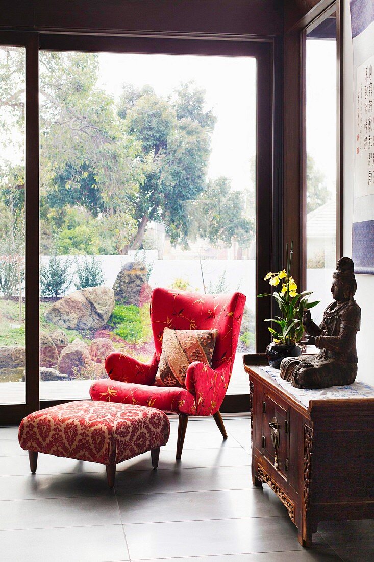 Eleganter roter Lesesessel und Fussschemel vor raumhoher Verglasung mit Gartenblick