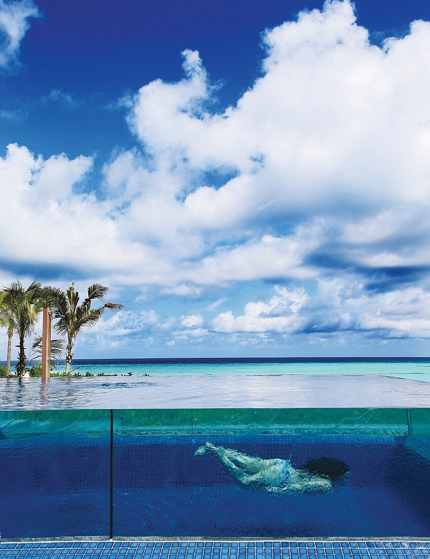 Frau taucht in Pool mit Glaswand, am Meer mit blauem Wolken-Himmel