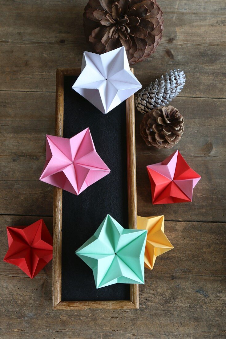 Colourful origami stars on chalkboard and pine cones on wooden surface