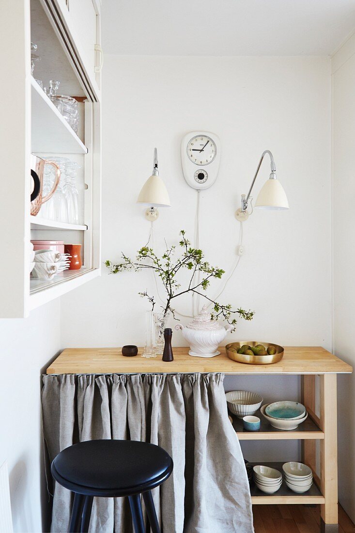 Holztisch mit Regalböden für Geschirr und hellgrauem Vorhang, darüber Wandleuchten und Retro Wanduhr