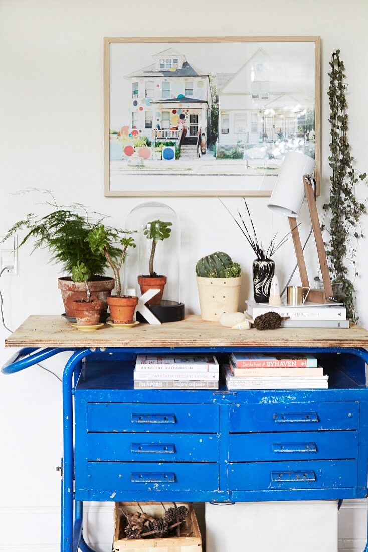 Blaue Vintage Werkbank mit Pflanzenarrangement und Tischlampe vor gerahmtem Bild