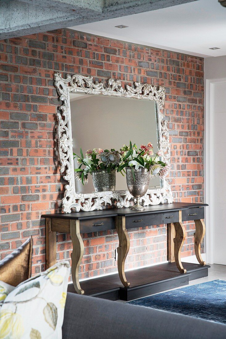 Kunsthandwerklicher Spiegel auf Konsolentisch an Ziegelwand gelehnt, davor Blumenstrauss in Silberkelch