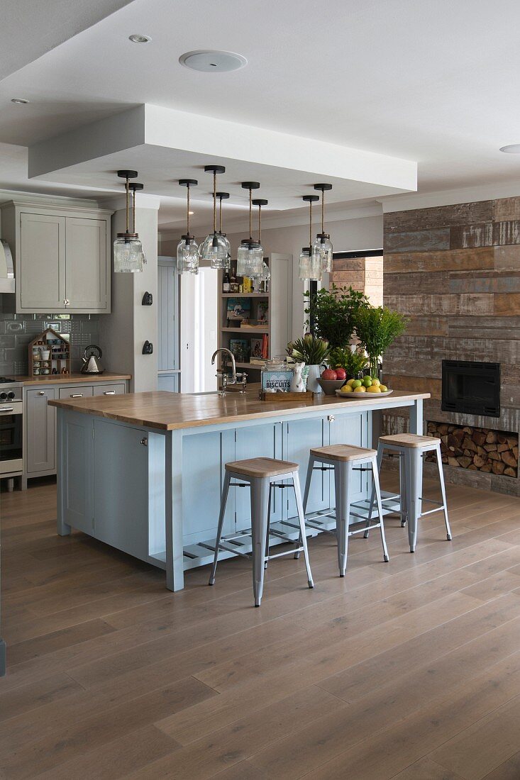 Dove-grey island counter and bar stools next to chimney breast with rustic, reclaimed-wood cladding in kitchen