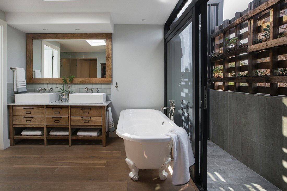 weiße, freistehende Badewanne vor offener Schiebefenstertür und Blick auf rustikales Holzbrüstungsgitter