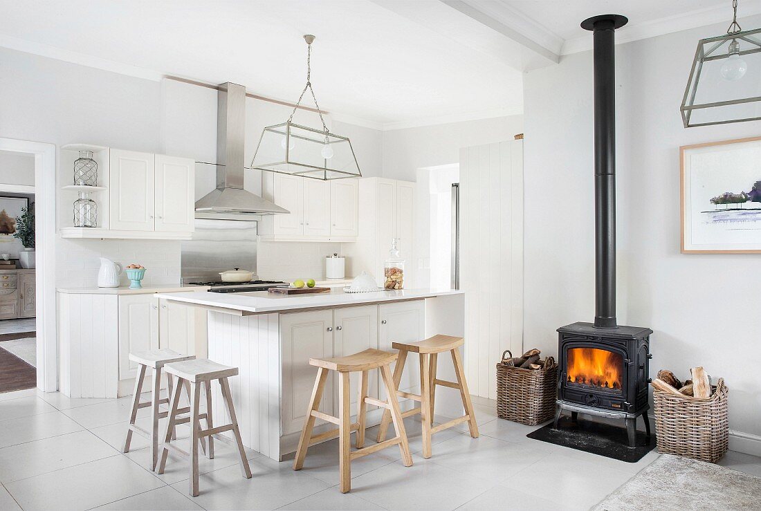 Offene, weiße Landhausküche mit schwarzem Schwedenofen und rustikalen Holz-Barhockern an Küchentheke