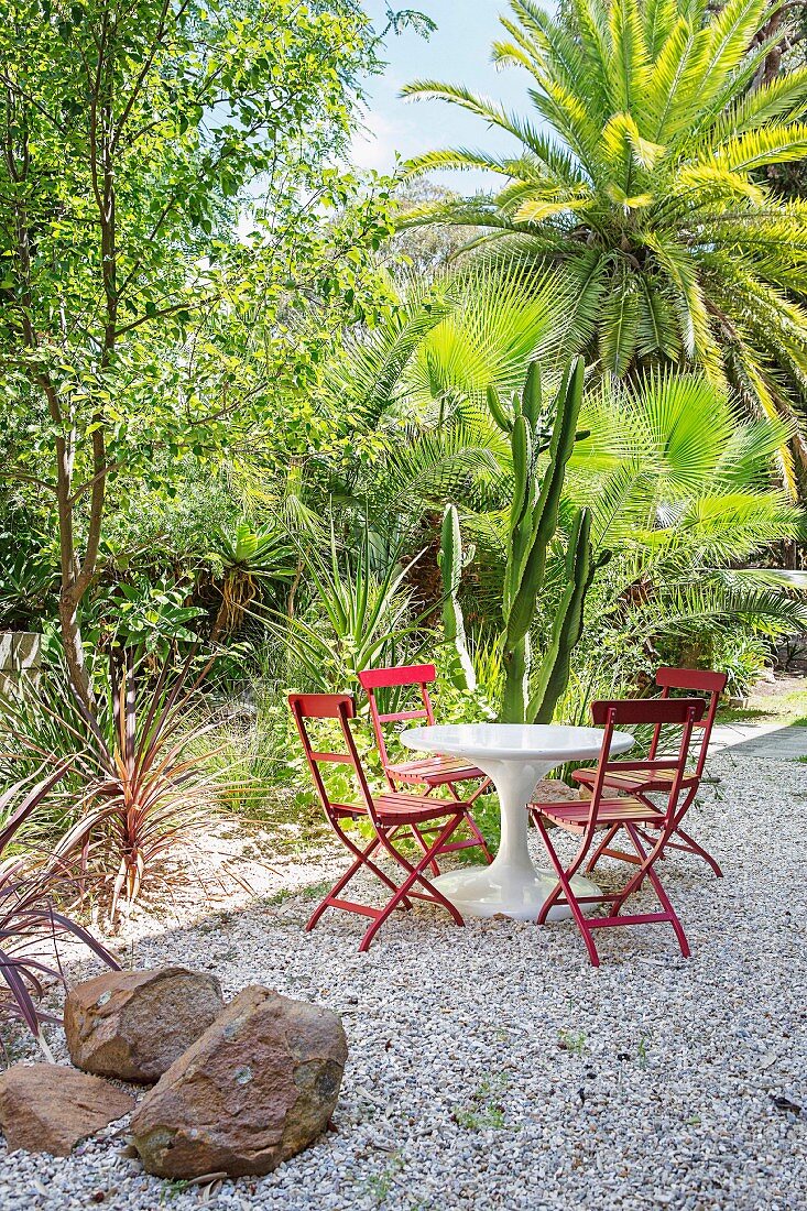 White table with red chairs on gravel in front of exotic plants