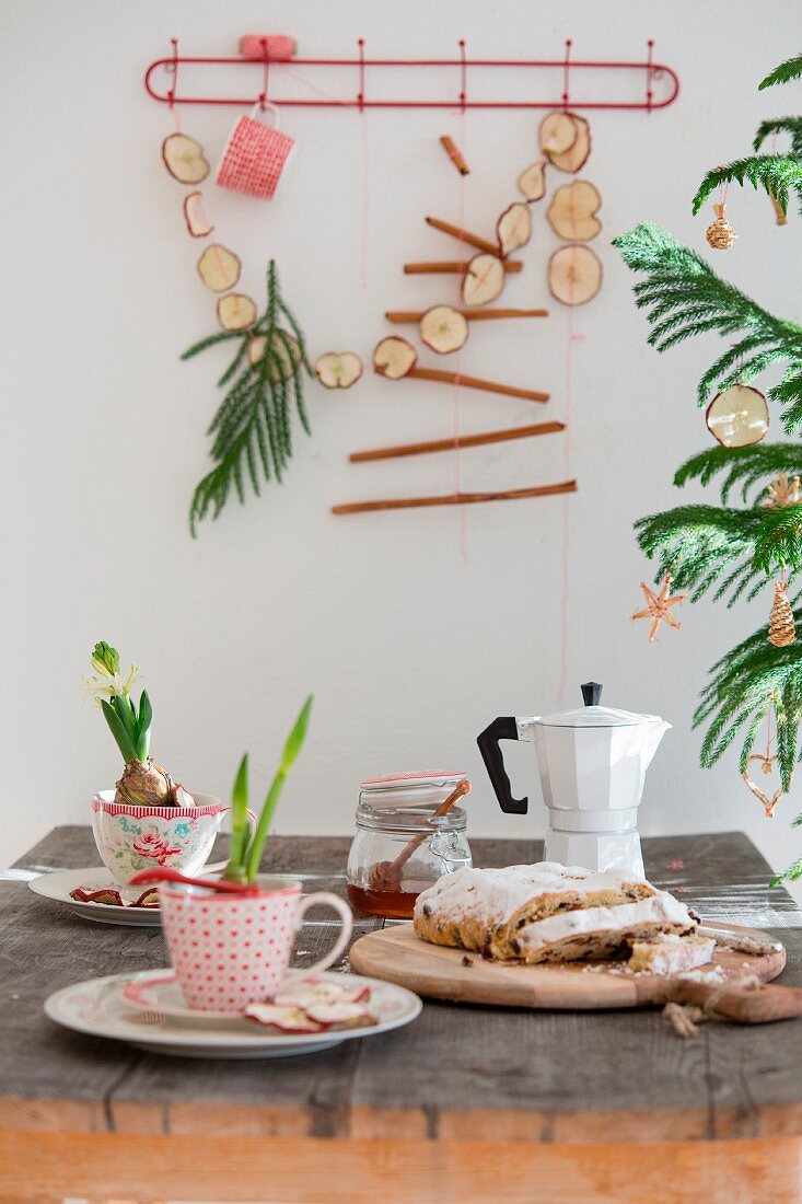 Weihnachtstisch mit Hyazinthe und Amaryllis in nostalgischen Tassen, Stollen und Espressokanne, im Hintergrund Girlande aus getrockneten Apfelscheiben und Zimtstangen an der Wand
