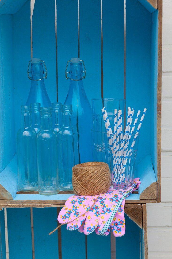 Aufbewahrungregal aus blau gestrichenen Holzkisten mit Flaschen, Garnrolle, Strohhalmen und Gartenhandschuhen