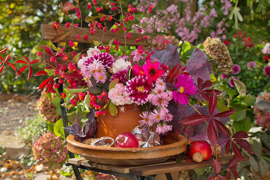 Herbststrauß mit Kohlblättern, Dahlien, Äpfeln, Walnüssen und Glasprismen auf Gartenstuhl im Garten