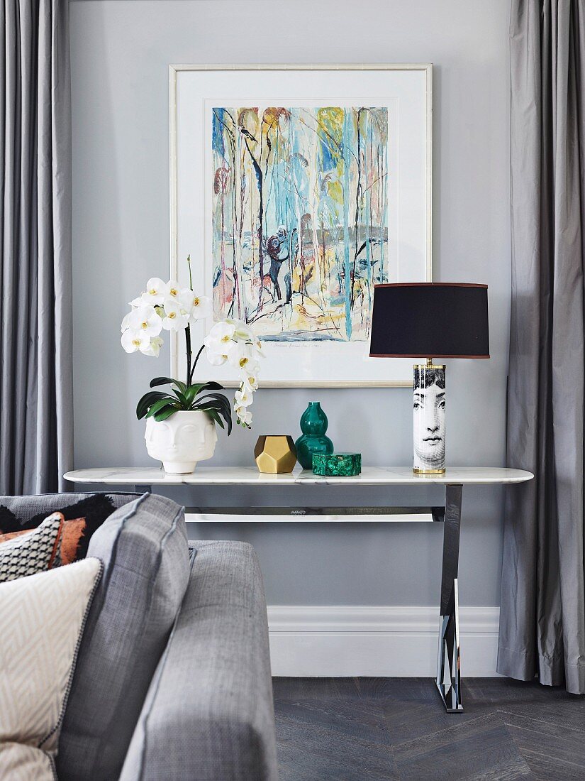 White orchid, vases and Fornasetti table lamp on console table in front of modern picture on gray wall