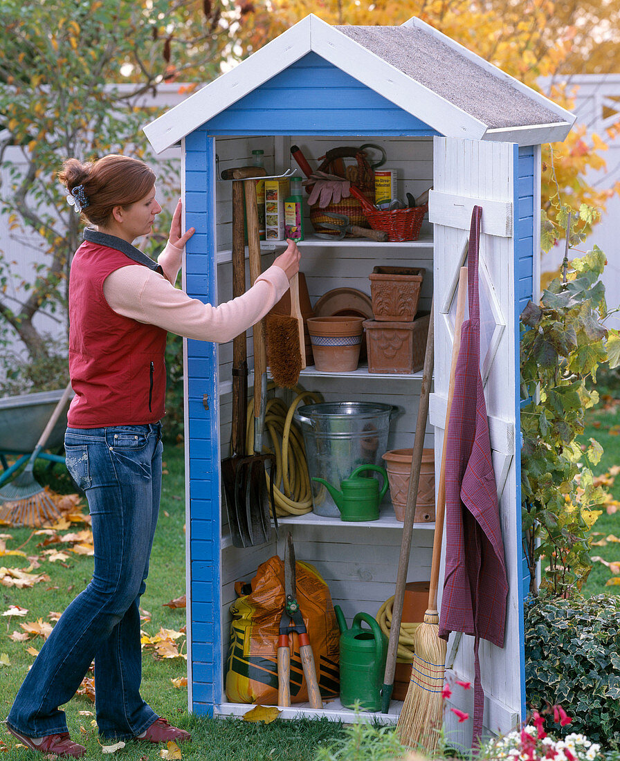 Frau hängt Besen in kleines Gerätehaus mit Töpfen und Gartengeräten