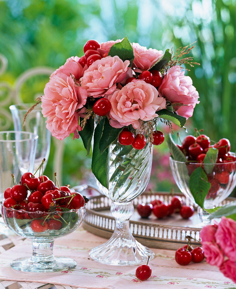 Strauß aus Rosa (Rosen), Prunus (Kirschen) und Gräsern in Glasbecher, Schale