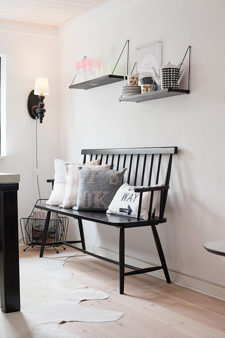 Black bench with scatter cushions below two shelves of crockery on wall
