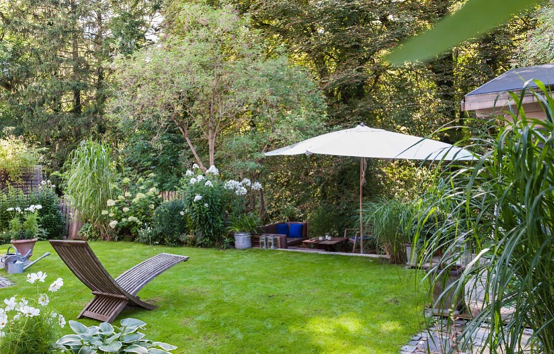 Holzliegestuhl auf Rasenfläche und Sonnenschirm in gepflegtem Garten