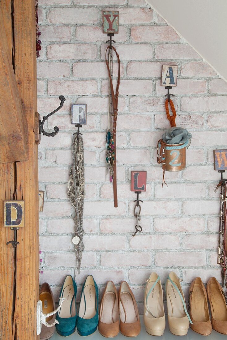 Various accessories and jewellery hung from vintage hooks on brick wall above collection of high-heeled shoes
